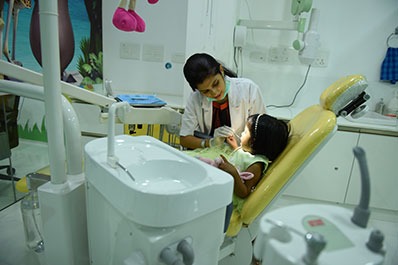 A female doctor sits on a chair and examines the child's oral hygiene with a torch beside her.