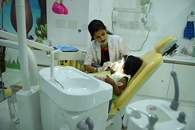 A female dentist sits on a chair & examines the child's oral hygiene with a torch beside her.